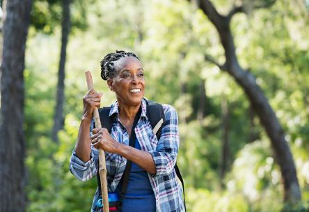 A senior African-American woman in her 70s hiking in a park smiling and leaning on a walking stick, standing with trees and lush foliage in the background.